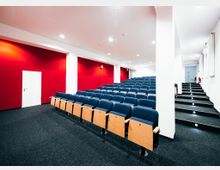 Ein moderner Hörsaal mit ansteigendem Sitzbereich, ausgestattet mit blauen Polstersitzen und hölzernen Klappschreibtischen. Die Raumgestaltung umfasst rote Wände, weiße Decken mit eingebauten Downlights und einen mit dunkelgrauem Teppich ausgelegten Boden.
