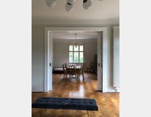 Ein Innenraum mit Parkettboden und hellen Wänden, durch eine geöffnete Schiebetür ist ein Essbereich sichtbar, der von einem Kronleuchter beleuchtet wird. Im Vordergrund steht eine gepolsterte Bank, und im Hintergrund befinden sich ein Esstisch mit Holzstühlen sowie ein Fenster mit Blick ins Grüne.