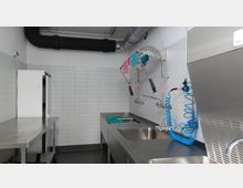 Industrielle Spülküche mit Edelstahloberflächen, mehreren Spülbecken und professionellen Sprühbrausen. Weiß geflieste Wände, grauer Boden und sichtbare Lüftungsrohre an der Decke prägen den Raum.