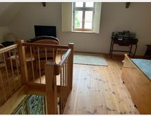 Heller Dachgeschossraum mit Holzboden und sichtbaren Holzbalken. Ein kleiner Bereich mit Rattanmöbeln, ein Fenster mit weißen Fensterläden, ein Tisch mit gestapelten Büchern und ein Geländer, das eine Treppe absichert, sind sichtbar.