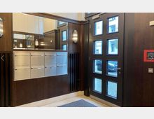 Eingangsbereich eines Wohngebäudes mit einer dunkelbraunen Holztür, die mehrere Glaseinsätze hat. Links an der Wand befinden sich silberne Briefkästen, darüber ein großer Spiegel und dekorative Wandleuchten.