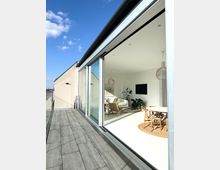 Moderne Dachterrassenwohnung mit großer Glasfront, die einen offenen Wohnraum mit Sofa, Stühlen, Pflanzen und einem Esstisch mit runder Teppichunterlage zeigt. Außenbereich mit grauem Fliesenboden und Blick auf städtische Gebäude im Hintergrund unter klarem Himmel.