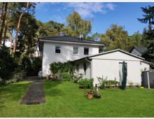 Zweistöckiges weißes Wohnhaus mit grauem Satteldach und angebautem Flachdachgebäude, umgeben von einem Garten mit Rasen, Topfpflanzen und Bäumen. Im Hintergrund sind hohe, bewaldete Kiefern und ein blauer Himmel zu sehen.