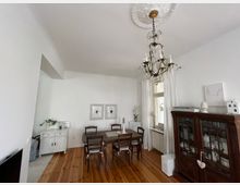 Esszimmer mit Holzboden, einem großen Kronleuchter, einem Esstisch mit Stühlen, einer weißen Kommode links und einem dunklen Vitrinenschrank rechts. Helle Wände mit zwei gerahmten Bildern, ein Fenster mit weißen Vorhängen und dekorativen Elementen.