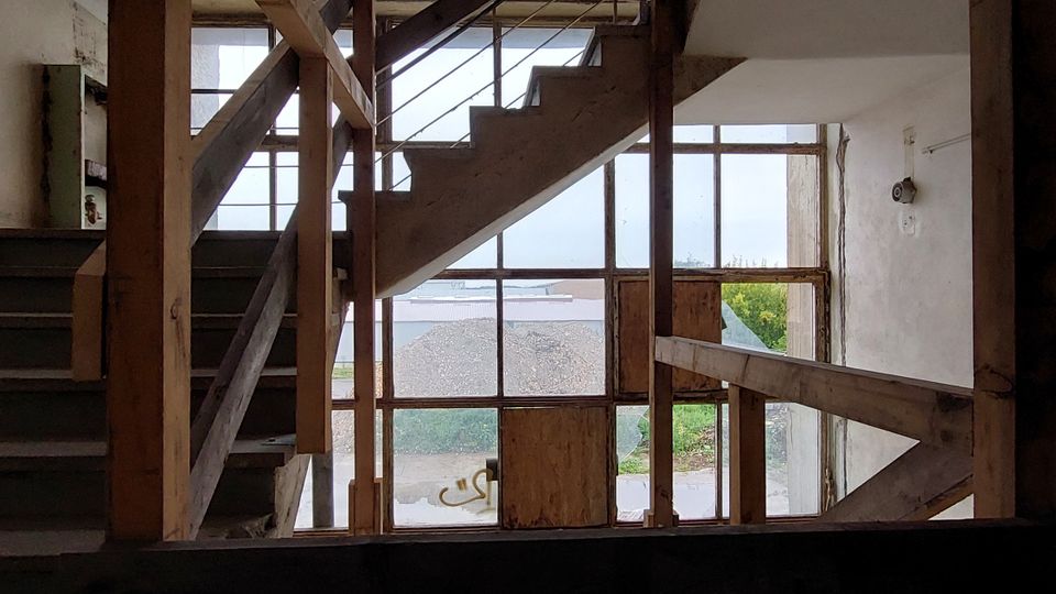 Treppenhaus in einem Rohbau mit freiliegendem Holz- und Metallgeländer, roh verputzten Wänden und großen Fenstern, die Tageslicht hineinlassen. Im Hintergrund sind Außenbereiche und Vegetation sichtbar.