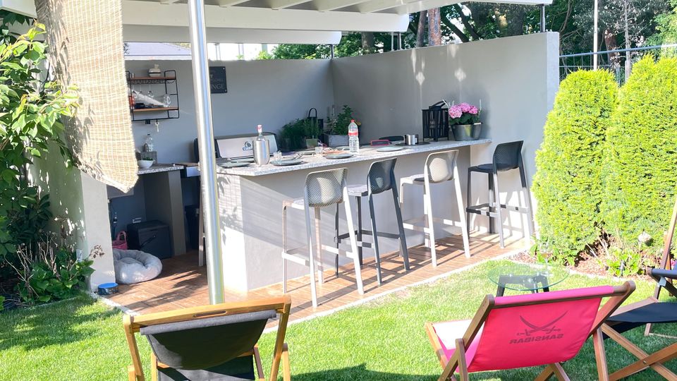 Moderne Gartenküche mit Überdachung, ausgestattet mit einer Theke, Barhockern und Küchenutensilien, umgeben von einer grünen Wiese und Bäumen im Hintergrund. Im Vordergrund stehen zwei Liegestühle auf dem Rasen.