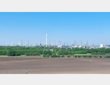 Weitläufige Industrielandschaft mit zahlreichen Schornsteinen und Windkraftanlagen am Horizont, umgeben von grünen Bäumen und Feldern im Vordergrund, unter klarem blauem Himmel.