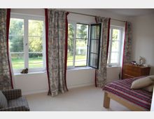 Schlafzimmer mit drei großen Fenstern, die mit gemusterten Vorhängen dekoriert sind, von denen eines geöffnet ist und einen Blick auf eine grüne Wiese und Bäume freigibt. Der Raum ist mit einem Holzbett mit gestreifter Bettwäsche, einer Holzkommode und einem karierten Sessel ausgestattet.