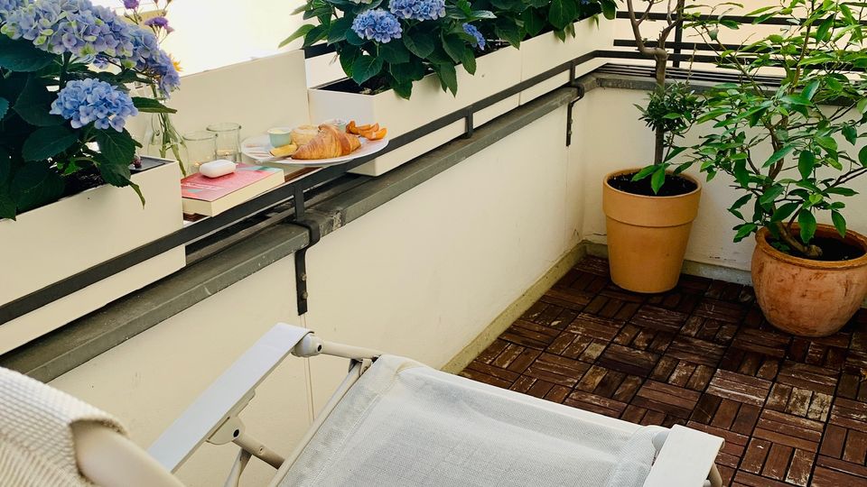 Kleiner Balkon mit Holzfliesen, weißer Liege und Geländer, auf dem mehrere Blumenkästen mit blauen Hortensien stehen. Im Hintergrund sind helle Hausfassaden mit Fenstern sowie zwei große Pflanzkübel mit Sträuchern zu sehen.