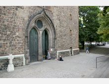 Außenseite einer historischen Kirche mit einer Fassade aus Natursteinmauerwerk und einem gotischen Portal mit dunkelgrünen Holztüren. Vor der Kirche ist ein Pflastersteinplatz zu sehen, ein kleines weißes Steinkreuz steht links, und mehrere Bäume begrenzen den Platz im Hintergrund.