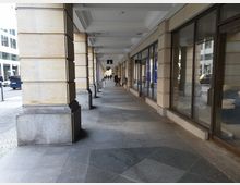 Überdachter Gehweg mit großen, rechteckigen Säulen aus hellem Stein auf der linken Seite und Schaufenstern auf der rechten Seite. Im Hintergrund sind Fußgänger sowie parkende und fahrende Fahrzeuge auf einer angrenzenden Straße zu sehen.
