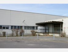 Ein Industriegebäude mit einer weißen Fassade, Fenstern im unteren Bereich und einem überdachten Ladebereich. Vor dem Gebäude befinden sich ein Metallzaun, Büsche und ein asphaltierter Bereich mit Straßenbegrenzungspfosten.