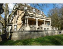 Historisches Herrenhaus mit heller Fassade, einem Walmdach und einem breiten, säulengestützten Portikus. Das Gebäude ist von Bäumen umgeben, und im Vordergrund liegt eine großzügige Steinbalustrade auf einem bepflanzten Rasen.