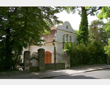 Großes Wohnhaus mit heller Fassade, geschwungenem Giebel und roten Dachziegeln, umgeben von dichtem Grün und hohen Bäumen. Im Vordergrund ist ein schmiedeeisernes Tor mit steinernen Pfosten zu sehen.