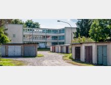 Mehrstöckiges, modernes Schulgebäude mit vielen Fenstern im Hintergrund, umgeben von Bäumen. Im Vordergrund eine Reihe von Garagen mit farbigen, teils verwitterten Holztoren und einem unbefestigten Weg, der durch das Gelände führt.