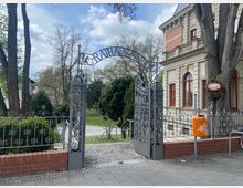 Eingangstor mit verschnörkeltem Metallzaun, beschriftet mit „Rathauspark“, führt in eine Grünanlage mit Bäumen und Gehwegen. Rechts im Bild steht ein historisches Gebäude mit verzierter Fassade und roten Fensterrahmen.