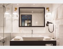 Modernes Badezimmer mit weiß gefliesten Wänden, einem rechteckigen Spiegel in schwarzem Rahmen und einem Waschbecken mit breiter Armatur. Links daneben befinden sich Glaslampen, rechts ein Wandfön, ein Handtuchhalter und gestapelte Handtücher.