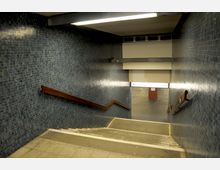 Treppenabgang in einer U-Bahn-Station mit blauen Kacheln an den Wänden, gelbem Beleuchtungselement an der Decke und einem hölzernen Handlauf. Unten ist der Bodenbereich der Station mit roten Wandkacheln erkennbar.