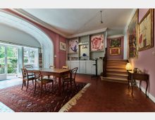 Wohnraum mit rosa Wänden, einer hölzernen Treppe und einer großen Fensterfront, die in einen Garten führt. Im Raum befinden sich ein Esstisch mit vier Stühlen, ein Teppich mit orientalischem Muster, Bücherregale und an den Wänden hängen verschiedene farbenfrohe Kunstwerke.