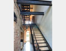 Industrie-inspirierter Innenraum mit freiliegenden Backsteinwänden, einer Metalltreppe mit offenen Stufen und einer Betonbodenfläche. Im Hintergrund ist eine Glastür zu sehen, durch die Tageslicht einfällt.