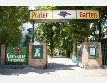 Eingang zum Pratergarten, einem Biergarten in Berlin, mit einem großen Schild über dem Tor und Hinweisschildern an Backsteinmauern. Im Hintergrund befindet sich ein von Bäumen gesäumter Weg.