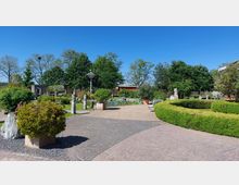 Ein gepflegter Garten mit gepflasterten Wegen, dekorativen Sträuchern in Töpfen und geschnittenen Hecken. Im Hintergrund befinden sich ein Teich mit umliegenden Bäumen und eine rote Gartenmauer.