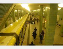 U-Bahnhof mit gelbem Zug an einem Bahnsteig, grüne Säulen und geflieste Wände dominieren das Erscheinungsbild. Zahlreiche Personen bewegen sich auf dem Bahnsteig, im Hintergrund ist eine Uhr und eine digitale Anzeigetafel sichtbar.