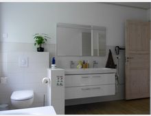 Ein modernes Badezimmer mit weißen Fliesenwänden, einem Hänge-WC und einem Waschbecken mit einer großen Spiegelfläche darüber. Eine Holztür, ein Handtuchhalter, und verschiedene Badezimmerartikel sind sichtbar; eine kleine Pflanze steht als Dekoration auf einem Vorsprung.