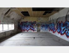 Ein verlassenes Gebäude mit beschädigtem Boden und Deckenplatten. Die Wände sind mit grauen, roten und blauen Graffiti bedeckt, und an der linken Seite befinden sich Fenster mit Blick nach draußen.