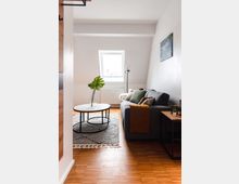 Wohnzimmer in einer Dachgeschosswohnung mit schräger Decke, einem zentralen Dachfenster, einem grauen Sofa mit dekorativen Kissen und einer gelben Decke, einem runden Couchtisch mit Glasvase auf einem grauen Teppich sowie Parkettboden.