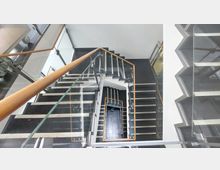 Ein modernes Treppenhaus in einem Gebäude mit schwarzen Stufen, weißen Kanten und Geländern aus Glas und Holz, das in mehreren Etagen spiralförmig nach unten führt. Die Wände sind weiß, und der Boden besteht aus dunklen Fliesen.