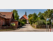 Straßenszene in einem Dorf mit einem gepflasterten Weg, der auf eine Kirche mit rotem Dach und Kirchturm zuführt, umgeben von Bäumen und Zäunen. Links ein Backsteinhaus mit Ziegeldach, rechts ein Straßenschild mit der Aufschrift „Kirchweg“.