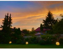 Ein Garten mit gepflegtem Rasen, blühenden Beeten und dekorativen Kugelleuchten im Vordergrund. Im Hintergrund sind hohe Tannenbäume, ein Holzzaun sowie Häuser mit roten und dunklen Dächern vor einem farbigen Himmel in Blau- und Orangetönen bei Sonnenuntergang zu sehen.