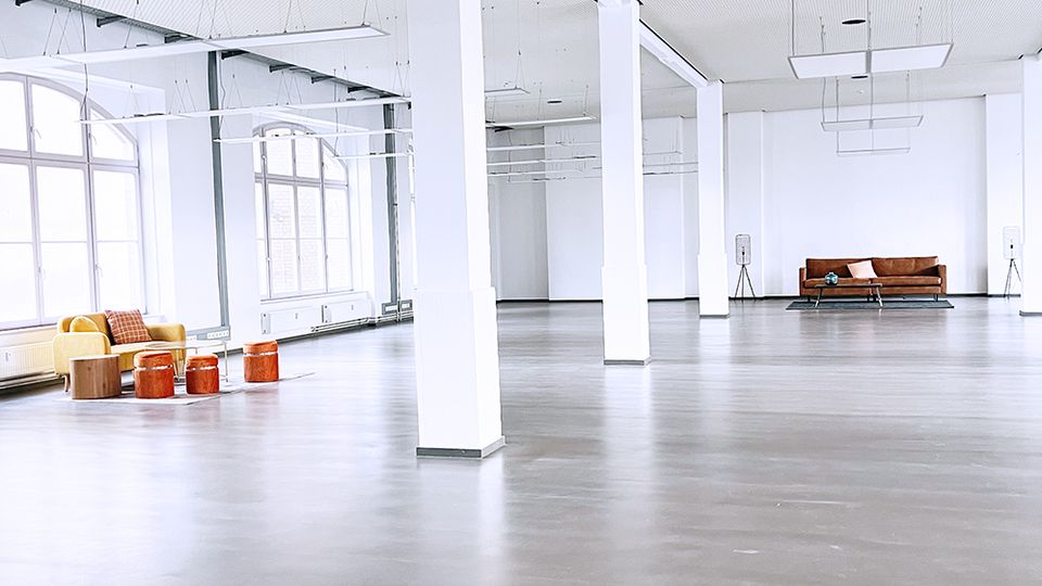 Großer, heller Raum mit glatten grauen Böden, weißen Wänden und mehreren weißen Säulen, die die Fläche gliedern. An den Seiten befinden sich vereinzelte Sitzmöbel wie ein gelbes Sofa und ein brauner Sessel.