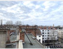 Blick von einem Flachdach mit roten Backsteinkaminen und Metallgeländern über eine Wohngegend mit mehrstöckigen Häusern mit roten und weißen Fassaden. Im Hintergrund sind kahle Bäume, parkende Autos und ein bewölkter Himmel zu sehen.