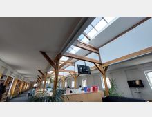 Großer, offener Leseraum mit sichtbaren Holzbalkenstrukturen und einem Oberlicht, das natürliches Licht einlässt. Der Raum ist mit Bücherregalen, großen Schreibtischen, Pflanzen und Sitzgelegenheiten ausgestattet.