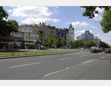 Straßenszene mit mehrspuriger Fahrbahn, flankiert von historischen und modernen Wohn- und Geschäftsgebäuden. Im Vordergrund eine U-Bahn-Station mit blauem Schild, Fußgänger und parkähnliche Grünstreifen; im Hintergrund Bäume und vereinzelte Fahrzeuge.