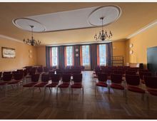 Ein Veranstaltungsraum mit Holzboden und orange gestrichenen Wänden, ausgestattet mit mehreren Reihen roter Stühle. Große Fenster mit Vorhängen lassen Tageslicht herein, während zwei Kronleuchter von der Decke hängen, und an den Wänden befinden sich dekorative Elemente sowie ein Gemälde.