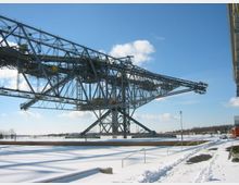 Großes Stahlgerüst eines Industriedrehkrans in schneebedeckter Landschaft, mit klar erkennbarem blauen Himmel und vereinzelt sichtbaren Wolken im Hintergrund. Im Vordergrund sind Wege und Geländer von Schnee bedeckt.