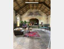 Großer Raum mit Holzbalkendecke und grobem Holzboden, eingerichtet mit einem Sofa, Stühlen, einem roten Teppich und einem Tischtennistisch. An der Rückwand befinden sich ein großer Rundbogen und verblasste Wandmalereien; durch breite Fenster fällt Licht herein.