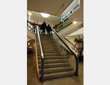 Treppenaufgang in einem Einkaufszentrum mit Glasgeländern und beleuchtetem Wegweiser mit der Aufschrift „Lift, Kasse“. Zwei Personen gehen die Treppe hinauf, umgeben von Pflanzen und Schaufenstern.