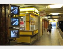U-Bahn-Station mit Verkaufsstand für Rostbratwurst, gelb verkleideter Kiosk mit Preistafel und Bildschirmen, die Bahnsteigansichten zeigen; im Hintergrund eine Uhr, Fahrplananzeige und Treppenaufgang.