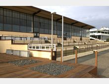 Große Tribüne mit gelber Fassade und einer Reihe von Fenstern, die den gesamten oberen Bereich bedecken. Davor befinden sich gestufte Sitzbereiche aus Beton mit Geländern, sowie ein Holzdeck mit eingelassenen Kiesflächen im Vordergrund.