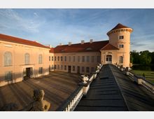 Barockschloss mit cremefarbenen Fassaden und roten Ziegeldächern, bestehend aus einem Hauptgebäude und einem runden Turm, umgeben von einem gepflasterten Hof und einer Balustrade. Im Hintergrund ist ein parkähnlicher Bereich mit Bäumen und einem See sichtbar.