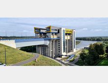 Modernes Schiffshebewerk mit grauer Betonfassade, blauen und gelben Akzenten. Es liegt an einem breiten Kanal, umgeben von grünen Wiesen, Bäumen und Gehwegen.