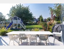 Garten mit einer Terrasse im Vordergrund, auf der ein weißer Tisch und Stühle stehen. Im Hintergrund ein Spielhaus mit blauer Rutsche, ein Klettergerüst, Rasen, Büsche und Bäume sowie Wohnhäuser am Horizont.