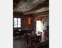 Gemütlicher Innenraum eines rustikalen Holzcafés mit Holztischen und -stühlen, einer Holzdecke und Wänden aus Holzbohlen. An der Wand hängen Bilder und dekorative Leuchten, und ein Fenster sowie eine geöffnete Tür lassen natürliches Licht herein.