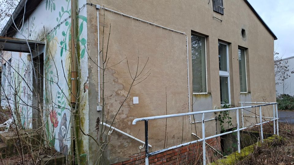Seitenansicht eines älteren, verputzten Gebäudes mit drei hohen Fenstern und einer kleinen Rampe mit weißem Metallgeländer entlang der Fassade. Links ist ein Wandgemälde mit Pflanzenmotiven zu sehen; davor stehen kahle Sträucher und überwucherte Bereiche am Sockel aus Ziegeln.
