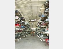 Ein großes Lager mit mehreren Regalen, auf denen gebrauchte Autos in mehreren Ebenen gestapelt sind. In der Mitte hebt ein Gabelstapler ein Fahrzeug an, und die Halle wird von einem Dach mit Lichtplatten überspannt, das natürliches Licht hereinlässt.