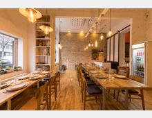 Gemütlicher Speiseraum mit hölzernen Esstischen und Stühlen, minimalistischer Einrichtung und einer Rückwand aus unverputztem Ziegelmauerwerk. Links ein großes Fenster mit Blick nach draußen, rechts ein Kühlschrank mit Getränken, und an der Decke hängen lange Reihen von Glühbirnenlampen.